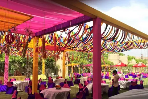 An outdoor party under a pink and yellow canopy strung with rainbow streamers and hanging lanterns, round tables and purple chairs set out on the lawn