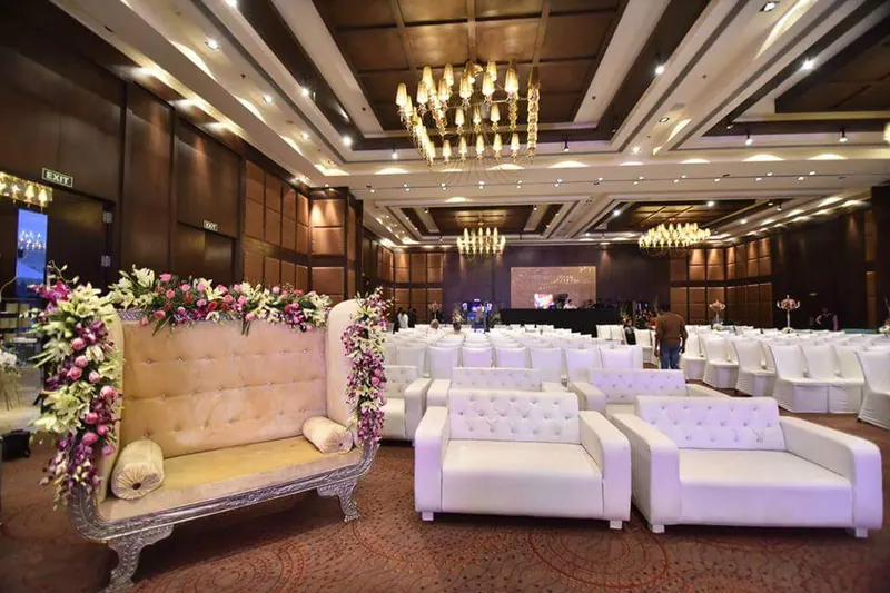 A reception stage of white tufted sofas behind a floral runner, under chandeliers in a wood-panelled hall