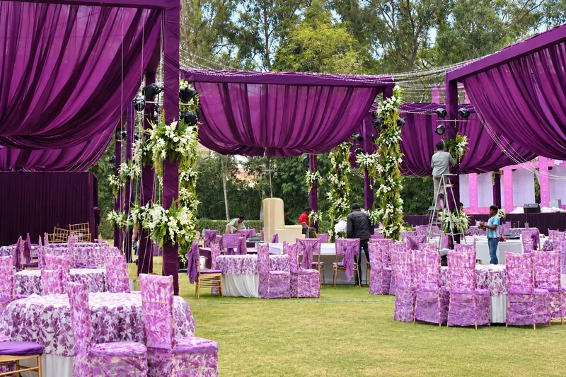 Outdoor setup with purple drapes and floral pillars, hanging white garlands above round tables in lilac-print covers