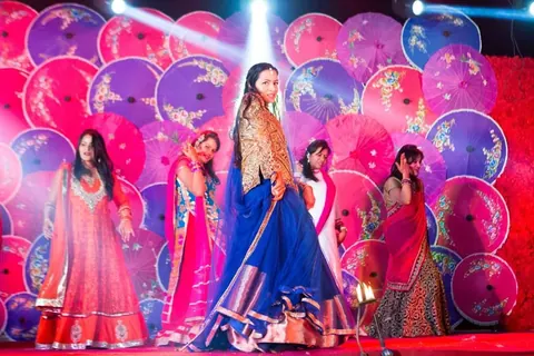 Women in lehengas and saris dancing on stage in front of a wall of painted pink and purple parasols