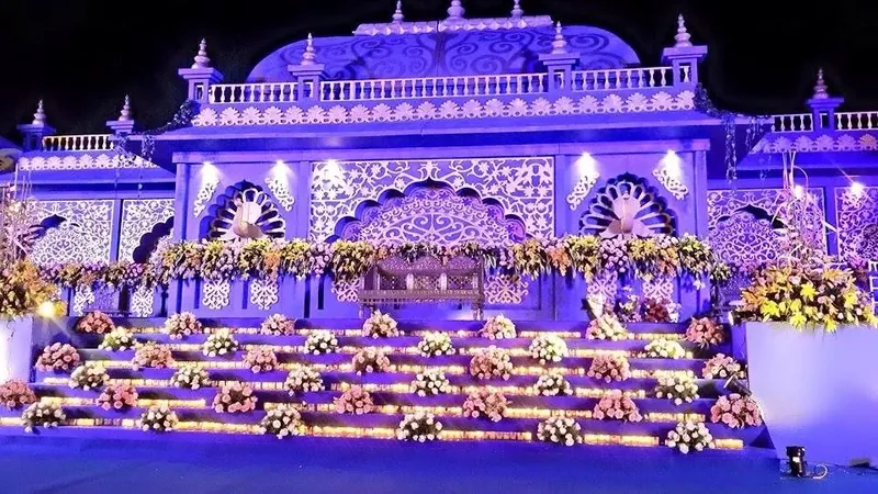 An illuminated wedding stage at night, its carved facade lit in lavender with tiered steps banked in flowers